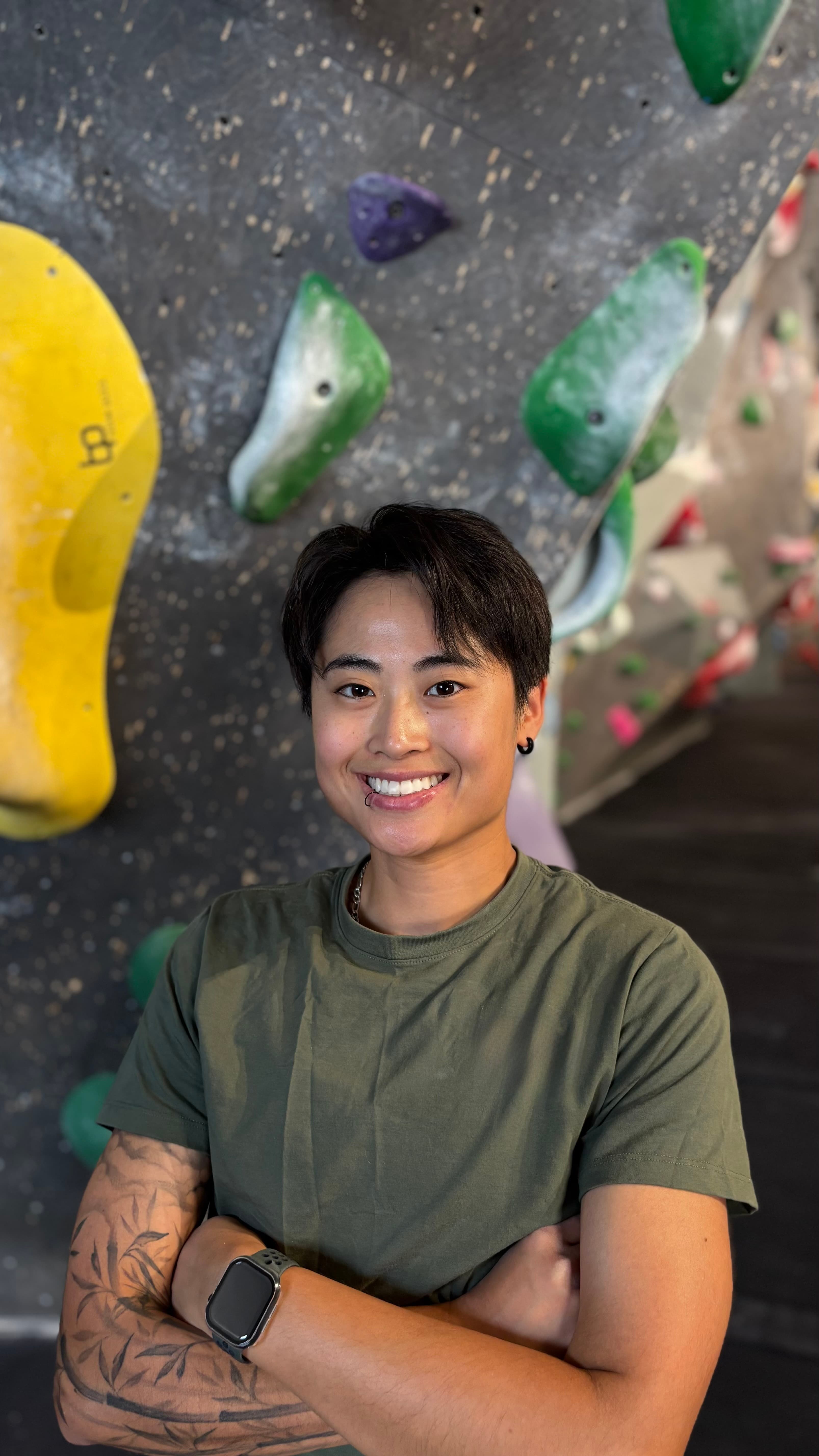 Smiling person with short hair and tattoos poses with crossed arms at a climbing gym.